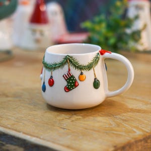 May include: White ceramic Christmas mug with a festive design. The mug features a green garland with colourful ornaments and a stocking. The handle is a classic D-shape. The mug is sitting on a wooden surface.