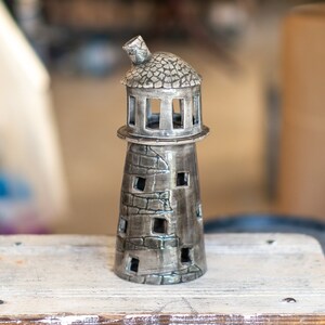 May include: A ceramic lighthouse figurine with a textured, gray finish. The lighthouse has a domed roof, rectangular windows, and a chimney. It is placed on a weathered, white wooden surface.