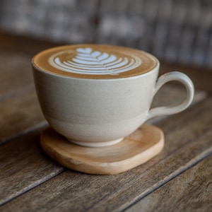 May include: A beige ceramic coffee cup filled with latte art, resting on a wooden coaster. The latte art features a leaf design. The cup has a curved handle and sits on a heart-shaped wooden coaster, all on a wooden surface.