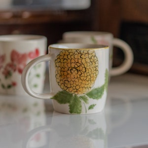 May include: White ceramic mug featuring a yellow floral design and green leaves. The mug has a white handle and a yellow rim. Other mugs with floral designs are visible in the background.