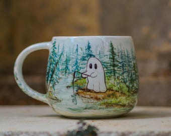 May include: White ceramic mug with a wraparound illustration of a ghost fishing in a forest scene. The design features a light blue river, green trees, and a cartoon ghost holding a fishing rod. The mug has a curved handle.