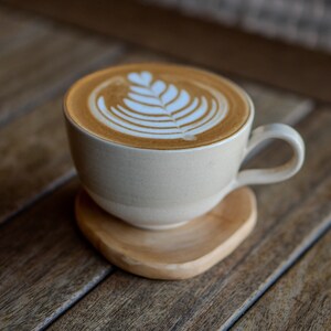 May include: A beige ceramic coffee cup with latte art, on a wooden coaster. The latte art shows a leaf design in the foam. The cup has a curved handle and sits on a wooden coaster. The coaster is made of wood.