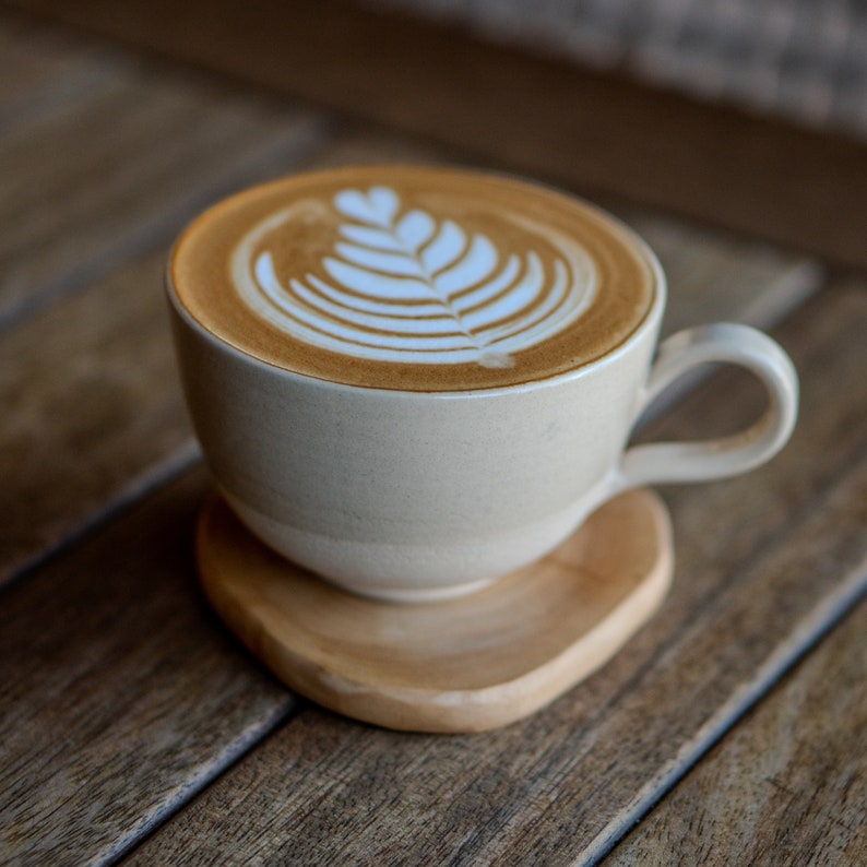 May include: A beige ceramic coffee cup with latte art, on a wooden coaster. The latte art shows a leaf design in the foam. The cup has a curved handle and sits on a wooden coaster. The coaster is made of wood.