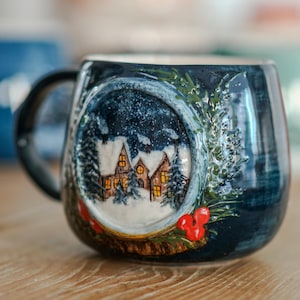 May include: A ceramic mug with a winter scene. The mug is dark blue with a handle and a detailed relief of a snowy village inside a circular frame. The frame is decorated with green foliage and red berries. The interior of the mug is white.