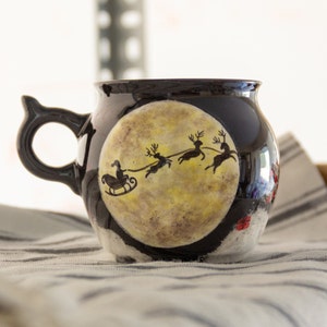 May include: Black ceramic mug with a whimsical design featuring Santa Claus in a sleigh pulled by reindeer against a full moon. The mug has a unique handle and is decorated with festive details, perfect for holiday beverages.
