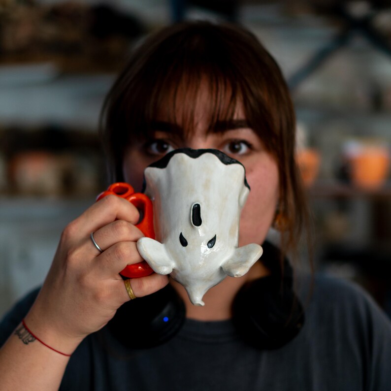 K&ouml;nnte beinhalten: Eine handgefertigte Keramik-Tasse in Form eines Geistes, mit wei&szlig;em K&ouml;rper, schwarzen Akzenten und einem roten Henkel. Die Tasse wird vor dem Gesicht gehalten, wobei die Augen der Person &uuml;ber der Tasse sichtbar sind. Die Tasse hat ein einzigartiges, skurriles Design.