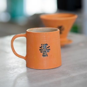 May include: Orange ceramic mug with a handle and a black and blue graphic design. A matching orange ceramic pour-over coffee maker is in the background. The mug has a cylindrical shape.