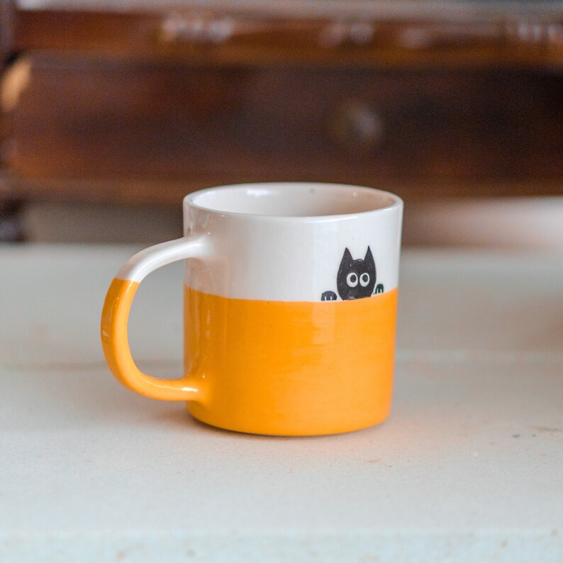 May include: A ceramic mug with a white upper section and a bright orange lower section. A black cat illustration peeks over the orange section. The handle is orange. This mug is perfect for coffee, tea, or any beverage.