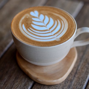 May include: A latte in a white ceramic mug with a leaf design in the foam, on a wooden coaster. The coffee is light brown, and the foam design is white. The mug has a handle and sits on a wooden coaster made of wood.