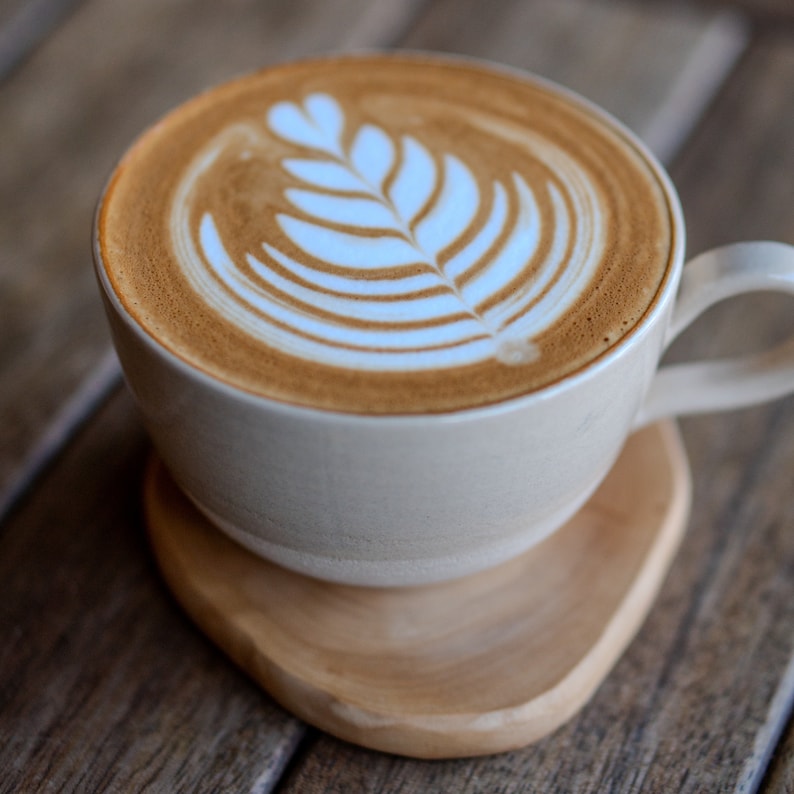 May include: A latte in a white ceramic mug with a leaf design in the foam, on a wooden coaster. The coffee is light brown, and the foam design is white. The mug has a handle and sits on a wooden coaster made of wood.