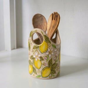 Pode incluir: Um porta-utensílios de cerâmica bege com um design de limão e floral. Contém uma colher e um garfo de madeira. O design apresenta limões amarelos, flores brancas e folhas verdes. O porta-utensílios tem uma alça.