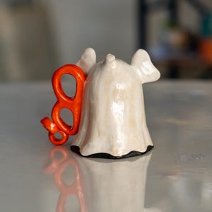 K&ouml;nnte beinhalten: Keramik-Geisterfigur mit einem orangefarbenen "B" daran. Der Geist ist cremefarben mit schwarzem Sockel und gl&auml;nzendem Finish. Das orangefarbene "B" ist ein dekoratives Element. F&uuml;r Halloween- oder gruselige Dekoration.