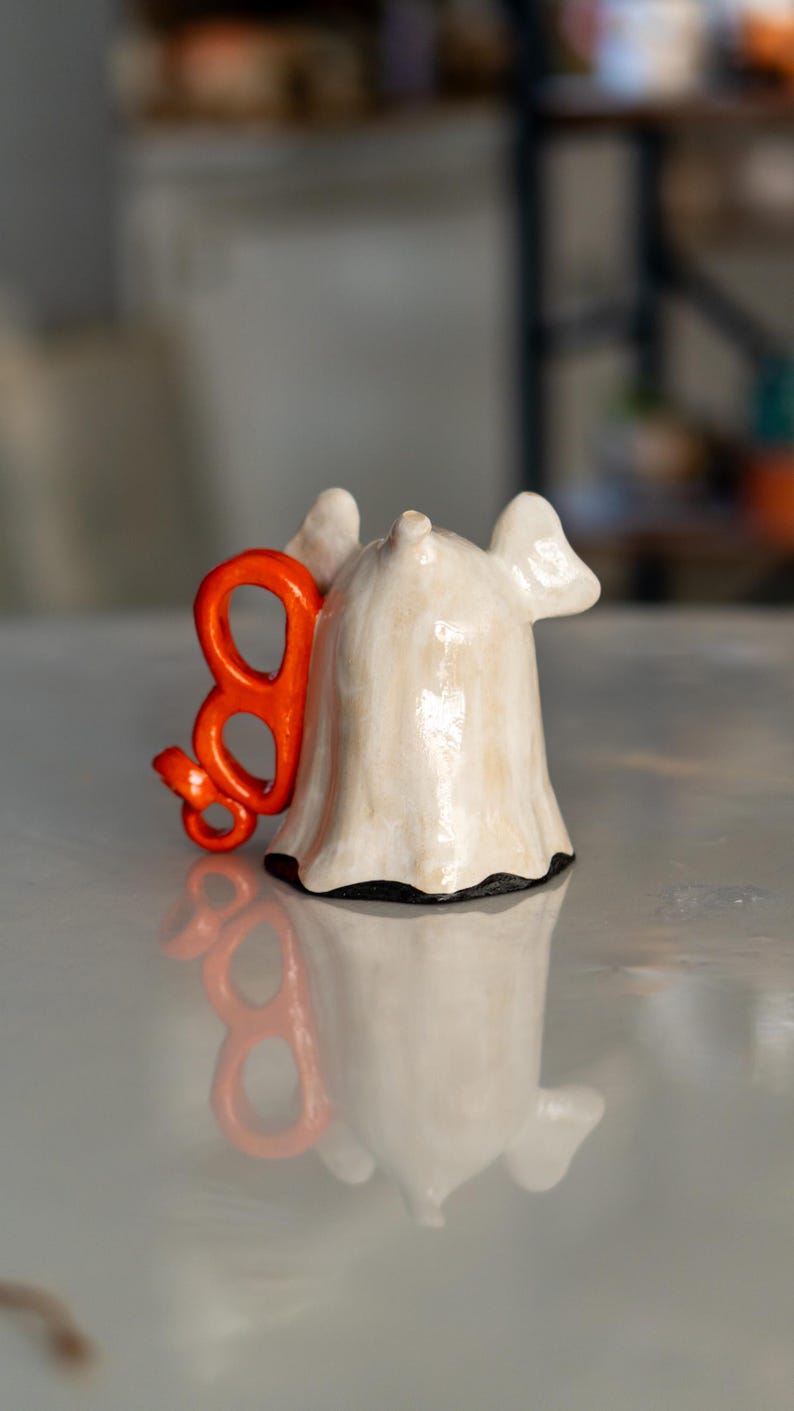 K&ouml;nnte beinhalten: Keramik-Geisterfigur mit einem orangefarbenen "B" daran. Der Geist ist cremefarben mit schwarzem Sockel und gl&auml;nzendem Finish. Das orangefarbene "B" ist ein dekoratives Element. F&uuml;r Halloween- oder gruselige Dekoration.
