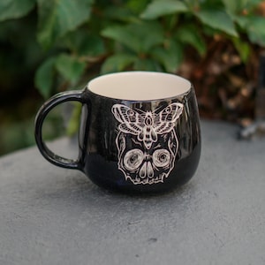 May include: Black ceramic mug with a white interior and a black handle. The mug features a white illustration of a moth over a skull. The mug is sitting on a grey surface.