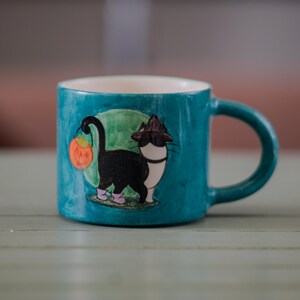 以下が含まれることがあります： 黒猫がカボチャを持っているイラストが描かれた、ターコイズ色のセラミックマグカップ。猫はピンクの靴と魔女の帽子をかぶっています。マグカップの内側は白く、持ち手はターコイズ色です。