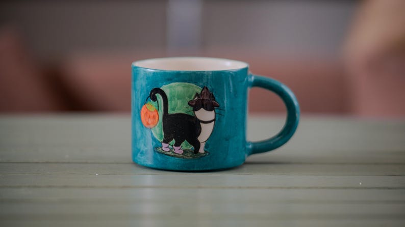 以下が含まれることがあります： 黒猫がカボチャを持っているイラストが描かれた、ターコイズ色のセラミックマグカップ。猫はピンクの靴と魔女の帽子をかぶっています。マグカップの内側は白く、持ち手はターコイズ色です。