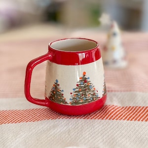 May include: A ceramic mug with a red handle and rim, featuring hand-painted Christmas trees. The mug has a cream-coloured body with colourful ornaments on the trees. The mug is approximately 10 cm tall.