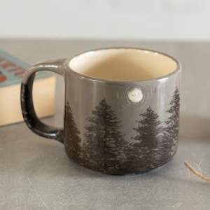 Puede incluir: Taza de cerámica gris con asa negra y escena de bosque. La taza presenta un fondo gris oscuro con árboles negros y una luna blanca. El interior de la taza es de color crema.