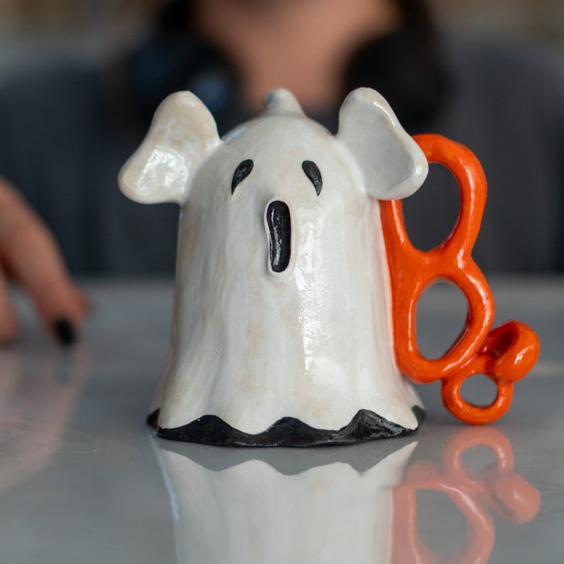K&ouml;nnte beinhalten: Eine wei&szlig;e Keramik-Geisterfigur mit schwarzen Gesichtsz&uuml;gen und einem orangefarbenen "Boo" daran befestigt. Der Geist hat einen weit ge&ouml;ffneten Mund und kleine Ohren. Halloween-Dekoration.