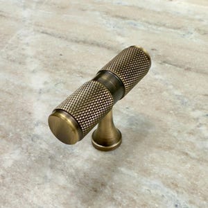 May include: A brass cabinet knob with a textured, cylindrical design. The knob has a T-shaped base.