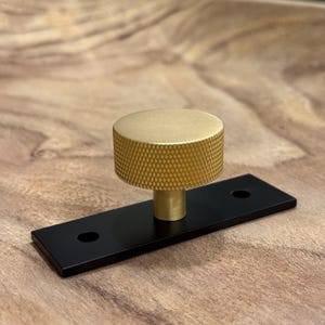 May include: A brass cabinet knob with a textured, cylindrical handle sits atop a black rectangular base. The hardware is designed for furniture and has a modern aesthetic. The knob is approximately 1.5 inches in diameter.
