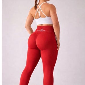 Puede incluir: Una mujer con un sujetador deportivo blanco y leggings rojos. Los leggings son de cintura alta y tienen el texto "N HIGH ATHLETICS" en la cintura. Lleva zapatillas deportivas negras.