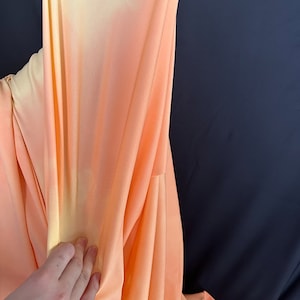 Könnte beinhalten: Ein peachfarbener und gelber Ombre-Stoff mit weicher, seidenartiger Textur. Der Stoff ist drapiert und gefaltet und zeigt seine glatte Oberfläche und den subtilen Farbverlauf.