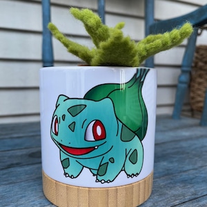 May include: A white ceramic planter with a green cartoon image of a Pokemon character, Bulbasaur, on the front. The planter has a bamboo base and is filled with a green succulent plant.