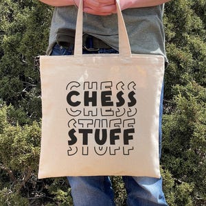 May include: A beige tote bag with the words "CHESS STUFF" in large, black, stylized letters. The bag has two handles and is held in front of a green, leafy background. The text is in a playful, rounded font.
