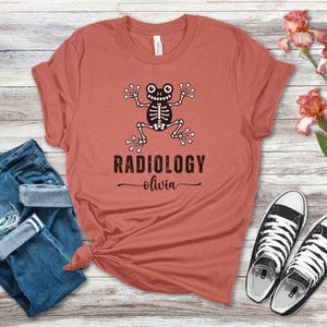 以下が含まれることがあります： 漫画のスケルトンデザインと「RADIOLOGY」の文字と「olivia」の名前が黒でプリントされたコーラルカラーのTシャツ。 シャツは青いジーンズと白と黒のスニーカーと組み合わせており、木製の表面に花が飾られています。