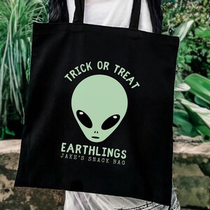 Może przedstawiać: Czarna torba z zielonym motywem kosmity i napisem "Trick or Treat" nad głową kosmity. Poniżej tekst "Earthlings" i "Jake's Snack Bag". Tło z zielonymi liśćmi.