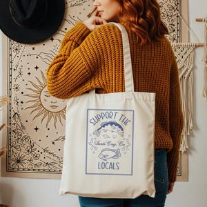 May include: Cream-colored tote bag with a blue graphic that reads "SUPPORT THE LOCALS" above illustrations of fish. The bag has a rectangular design with a blue border and the text "Santa Cruz, CA". The bag is held by a person wearing a mustard yellow sweater.