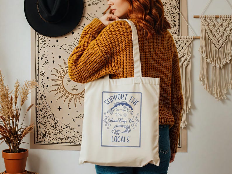 May include: Cream-colored tote bag with a blue graphic that reads "SUPPORT THE LOCALS" above illustrations of fish. The bag has a rectangular design with a blue border and the text "Santa Cruz, CA". The bag is held by a person wearing a mustard yellow sweater.