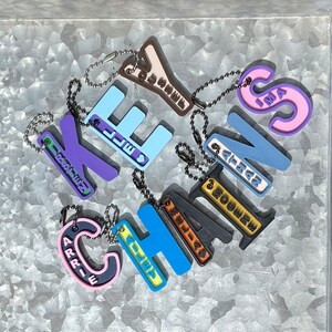 May include: A collection of colorful keychains with different names on them, including "Keychains", "Arrie", "Alley", "Charlie", "Ethan", "Boomer", "Ellie", "Kylie", "Emma", and "Beatriz".