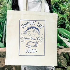May include: Cream-colored tote bag with a blue rectangular design featuring the text "SUPPORT THE LOCALS" and illustrations of fish, shells, and bubbles. The text "Santa Cruz, CA" is also included. The bag is held by a person.