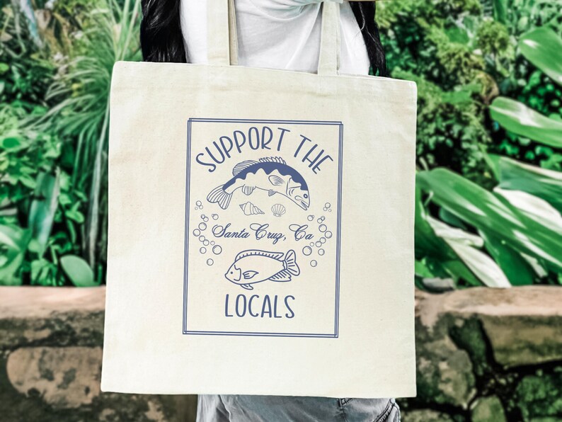 May include: Cream-colored tote bag with a blue rectangular design featuring the text "SUPPORT THE LOCALS" and illustrations of fish, shells, and bubbles. The text "Santa Cruz, CA" is also included. The bag is held by a person.