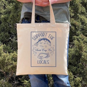 May include: Beige canvas tote bag with blue graphic text and illustrations. The text reads "SUPPORT THE LOCALS" with images of fish and bubbles. Below the text is "Santa Cruz, CA". The bag has two handles and is held by a person.