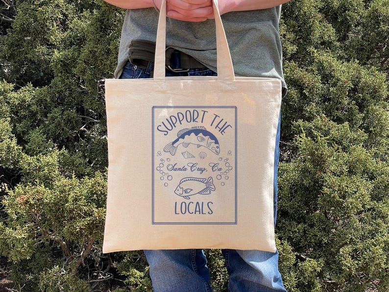 May include: Beige canvas tote bag with blue graphic text and illustrations. The text reads "SUPPORT THE LOCALS" with images of fish and bubbles. Below the text is "Santa Cruz, CA". The bag has two handles and is held by a person.