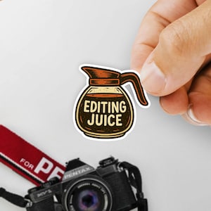 May include: A sticker featuring a coffee pot design with the words "EDITING JUICE" in a vintage style. The sticker is brown and tan with a black outline. A camera strap and camera are visible in the background.