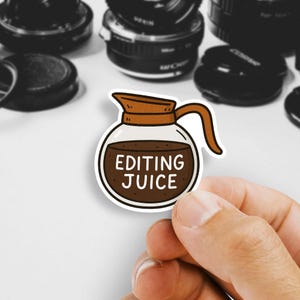 May include: A sticker featuring a cartoon coffee pot design with the words "EDITING JUICE" in a white font. The coffee pot is brown and filled with a dark brown liquid. The sticker is held by a hand, with camera lenses in the background.