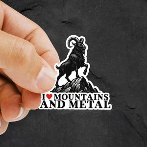 May include: A black and white sticker featuring a mountain goat standing on a rocky peak. The sticker has the text "I ❤️ MOUNTAINS AND METAL" in white, with a red heart symbol. The goat has large curved horns.