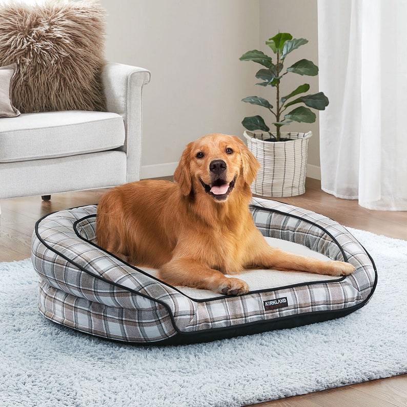 Peut inclure: Un lit pour chien en plaid brun et blanc avec une bordure noire. Le lit a un bord sur&eacute;lev&eacute; et une doublure douce et moelleuse. Un chien golden retriever est allong&eacute; sur le lit, regardant la cam&eacute;ra et souriant. Le lit porte le logo Kirkland Signature sur le c&ocirc;t&eacute;.