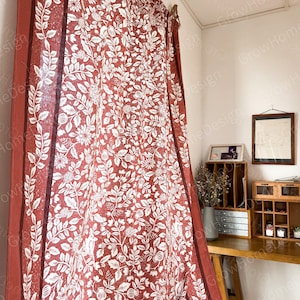 以下が含まれることがあります： 赤と白のフローラル柄のカーテンパネルで、茶色の縁取りが施されています。カーテンは、木製の棚と額縁に入った紙のある部屋にかけられています。