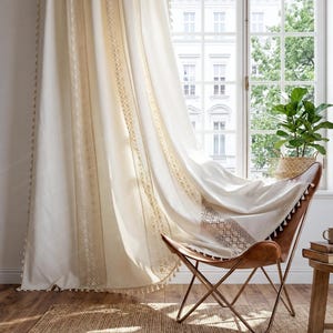 May include: Cream-colored curtains with intricate embroidered borders and tassel trim. A brown leather butterfly chair is draped with a matching cream throw, creating a cozy, bohemian-inspired interior design. A woven rug and potted plant complete the scene.