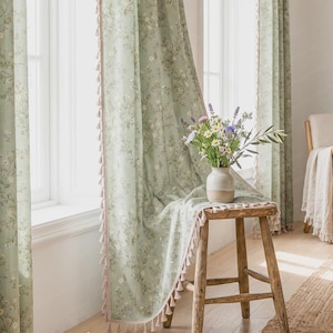 Può includere: Tende verde salvia con un delicato motivo floreale bianco e giallo e piccole nappe beige lungo i bordi. Un vaso in ceramica riempito con un bouquet di fiori di campo poggia su uno sgabello in legno drappeggiato con il tessuto stampato.