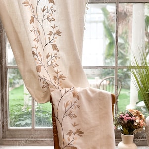May include: Beige linen curtain with embroidered brown floral design. The curtain features a botanical pattern along the edge and a tassel trim. The curtain is draped over a wooden chair in front of a window.