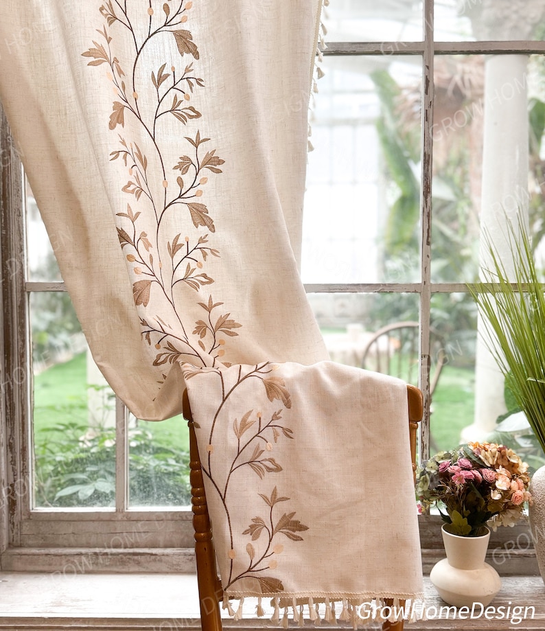 May include: Beige linen curtain with embroidered brown floral design. The curtain features a botanical pattern along the edge and a tassel trim. The curtain is draped over a wooden chair in front of a window.
