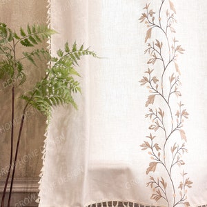 May include: Off-white linen curtain with a decorative embroidered floral design in shades of brown. The curtain has a fringe trim along the bottom edge. A green fern plant is visible on the left side of the image.