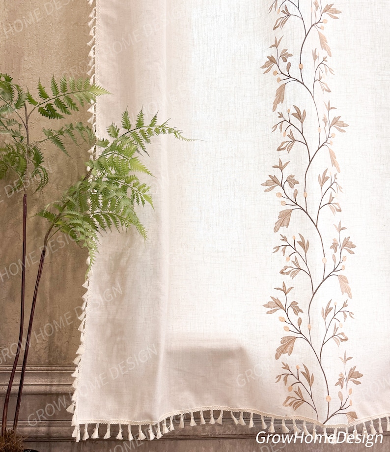 May include: Off-white linen curtain with a decorative embroidered floral design in shades of brown. The curtain has a fringe trim along the bottom edge. A green fern plant is visible on the left side of the image.