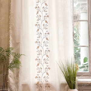 May include: Beige linen curtain with a vertical embroidered floral design in brown and tan. The curtain has a tassel trim along the bottom edge and a matching trim on the left side. The curtain hangs in front of a window.